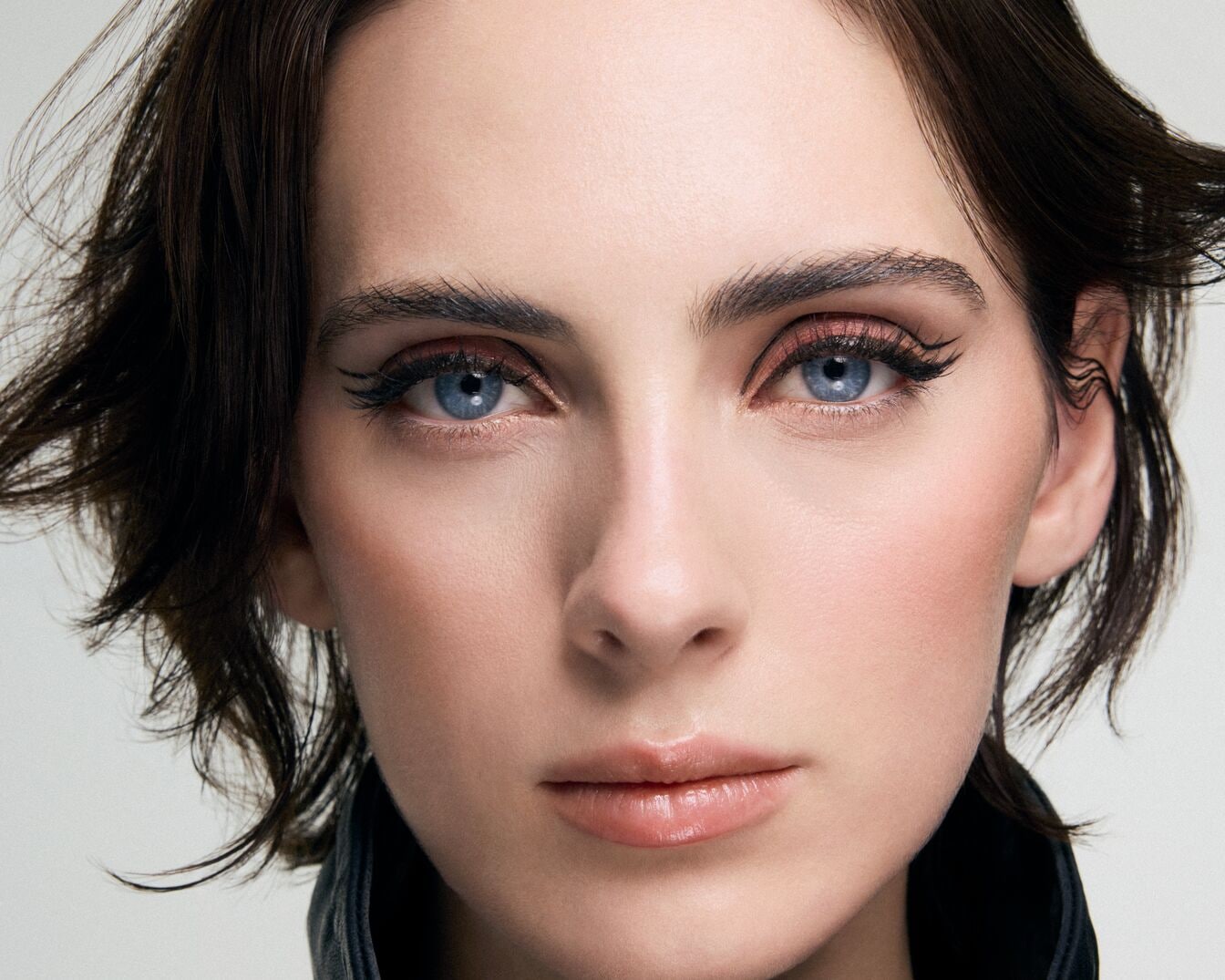 Nahaufnahme eines Frauengesichts mit hellblauen Augen, betont durch schwarzen Eyeliner und zarte Lippen, umrahmt von dunklem, gewelltem Haar.