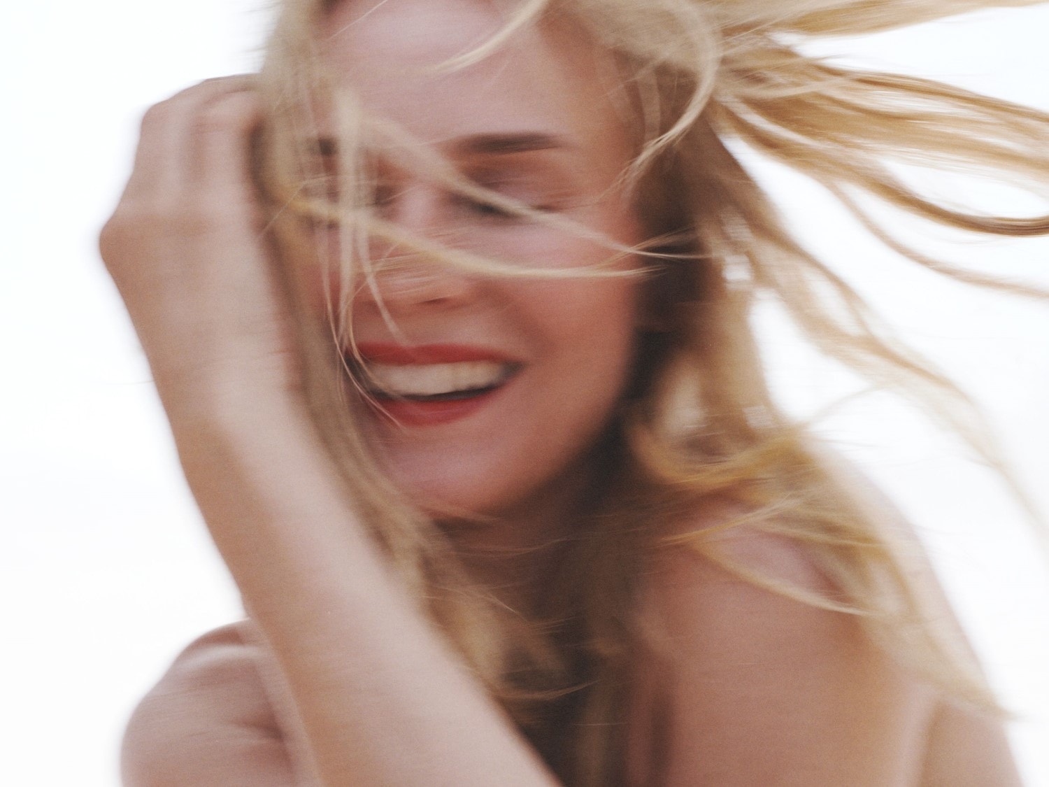 Nahaufnahme einer blonden Frau mit roten Lippen, die lacht und ihr Haar mit der Hand zurückwirft, während es im Wind weht.