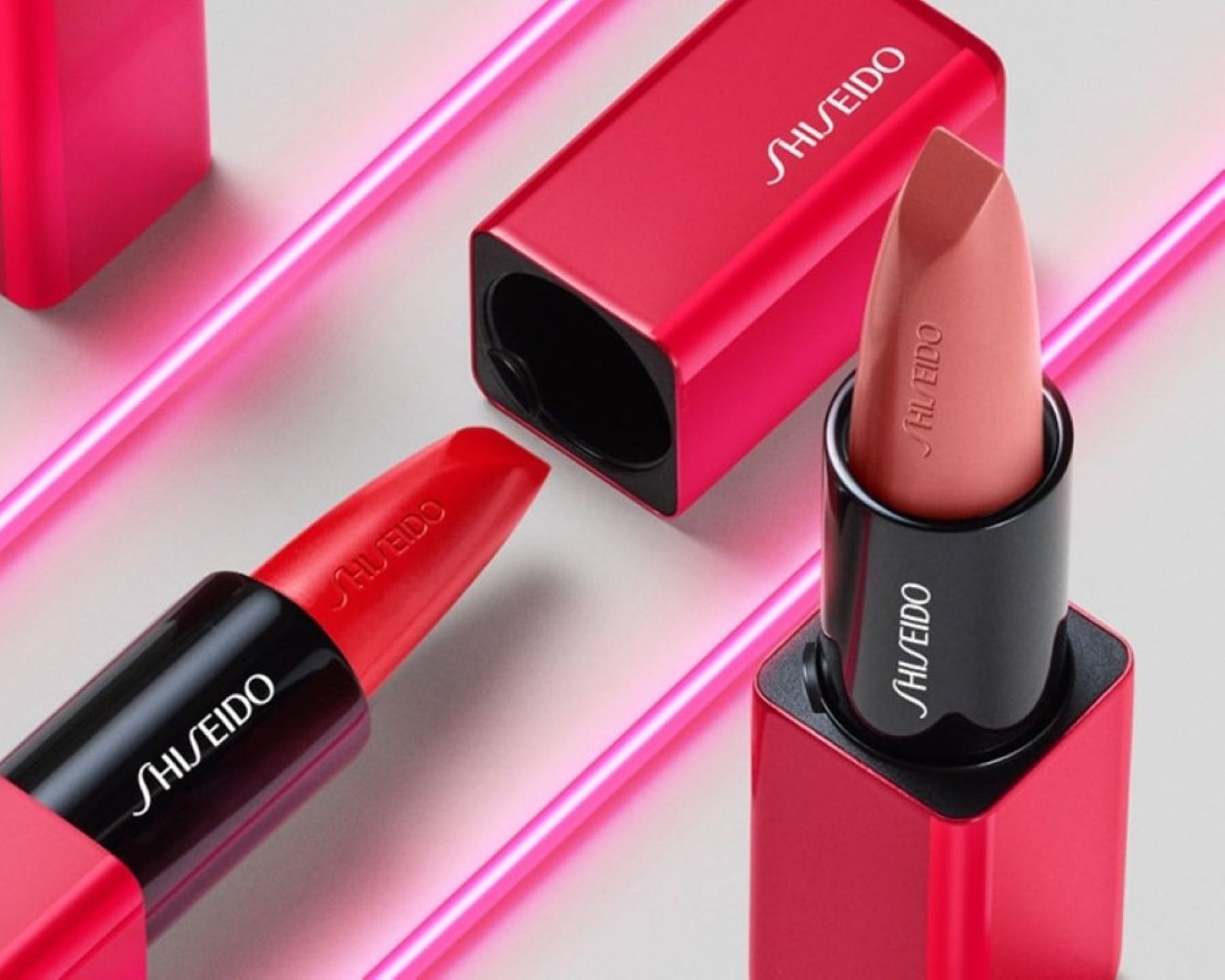 Nahaufnahme von zwei Shiseido-Lippenstiften: Einer ist rot mit schwarzer Basis, der andere ist pfirsichfarben mit roter Verpackung, beide liegen auf einem hellen Untergrund mit rosa Neonlinien.