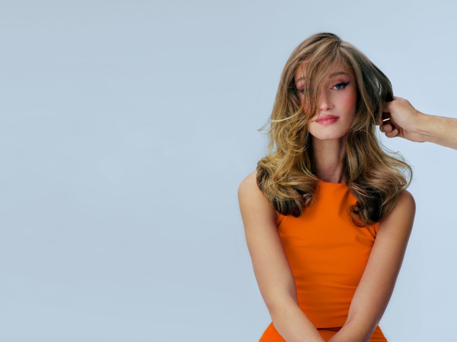 Een jonge vrouw met golvend bruin haar en blauwe ogen draagt een oranje top, terwijl een hand haar haar aanraakt.