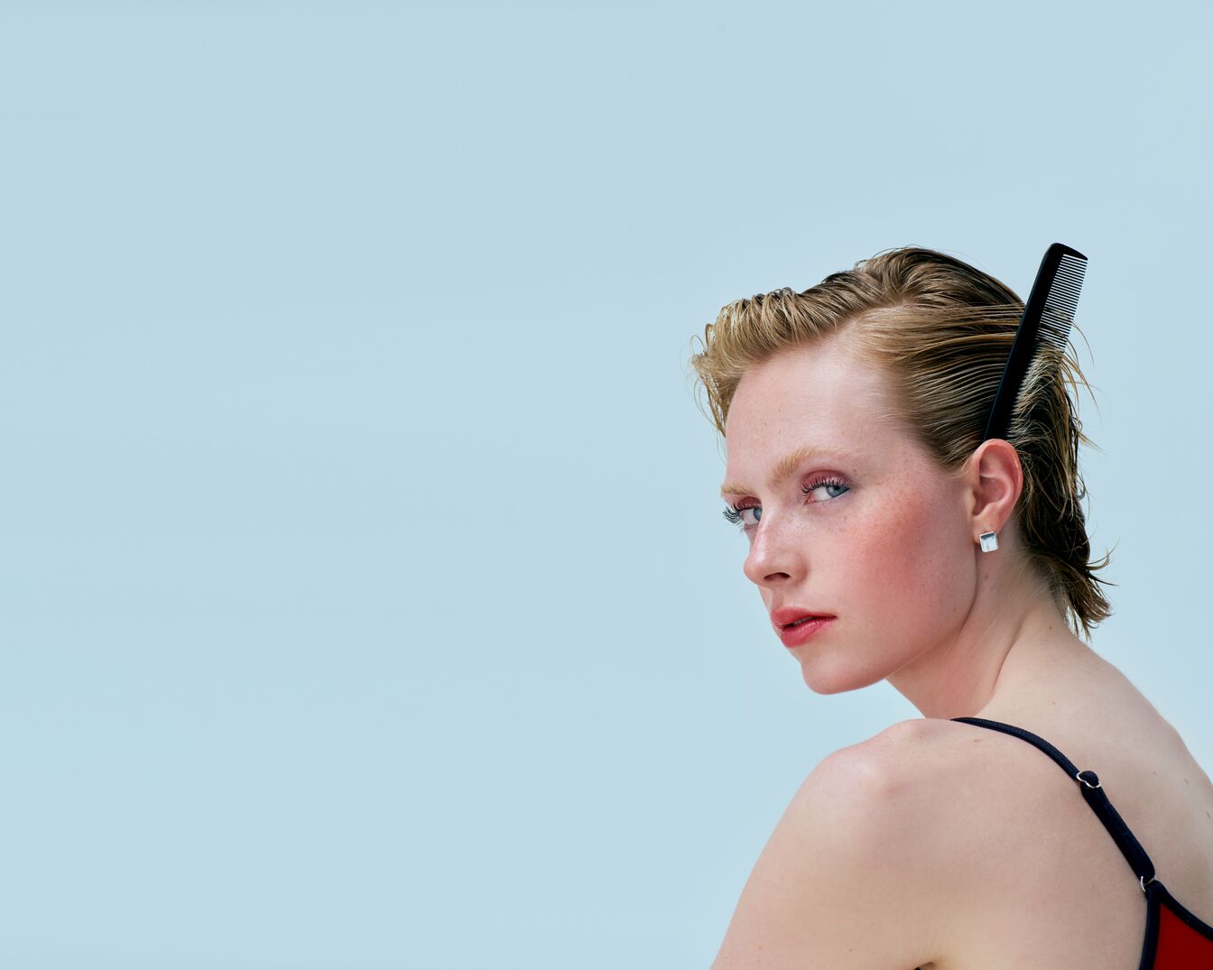 Een vrouw met kort, blond haar en een zwarte kam erin, kijkt over haar schouder met rode lippenstift en blush tegen een lichtblauwe achtergrond.