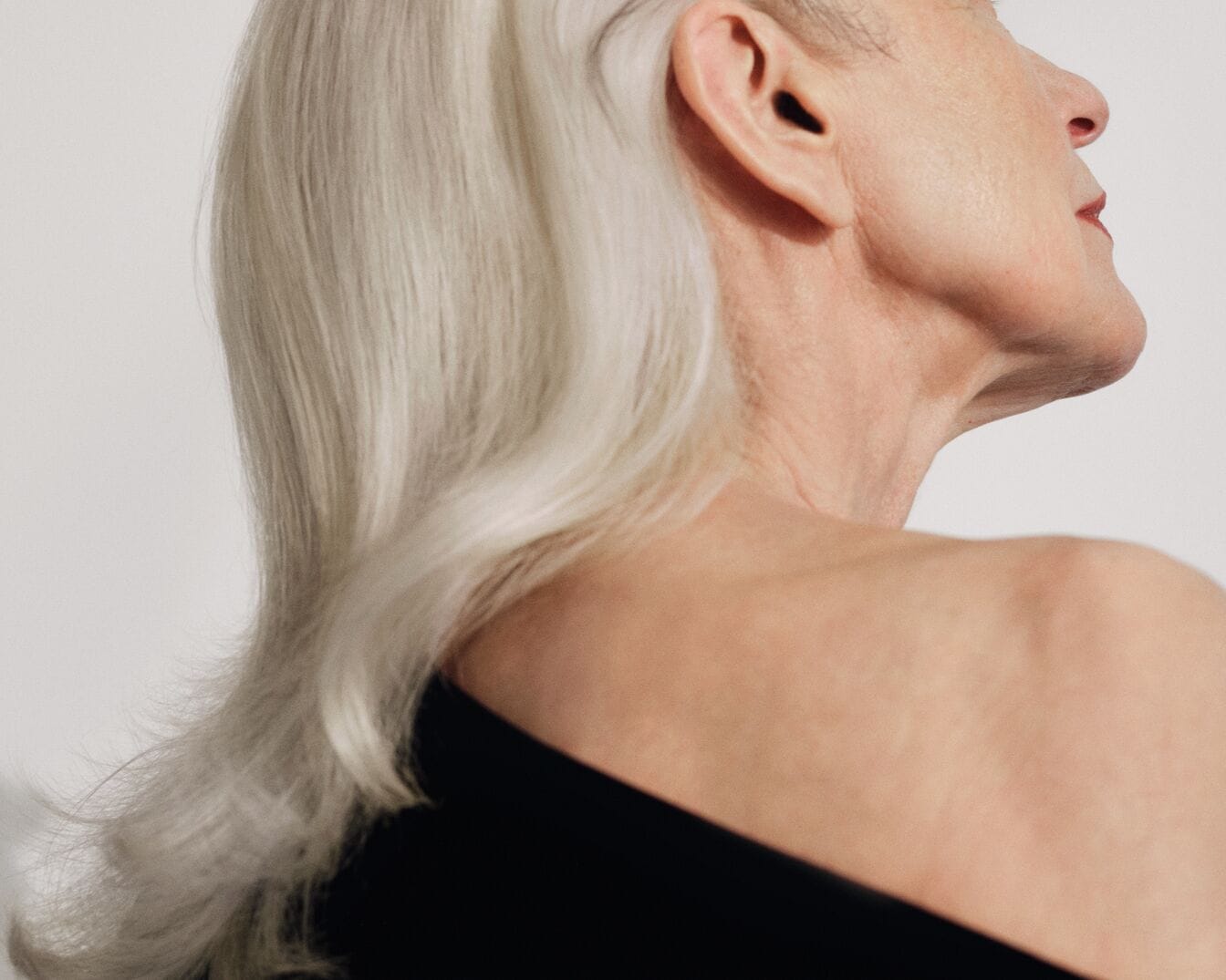Een vrouw met golvend, wit haar en een zwarte top, gefotografeerd van de zijkant, kijkend naar iets buiten beeld.