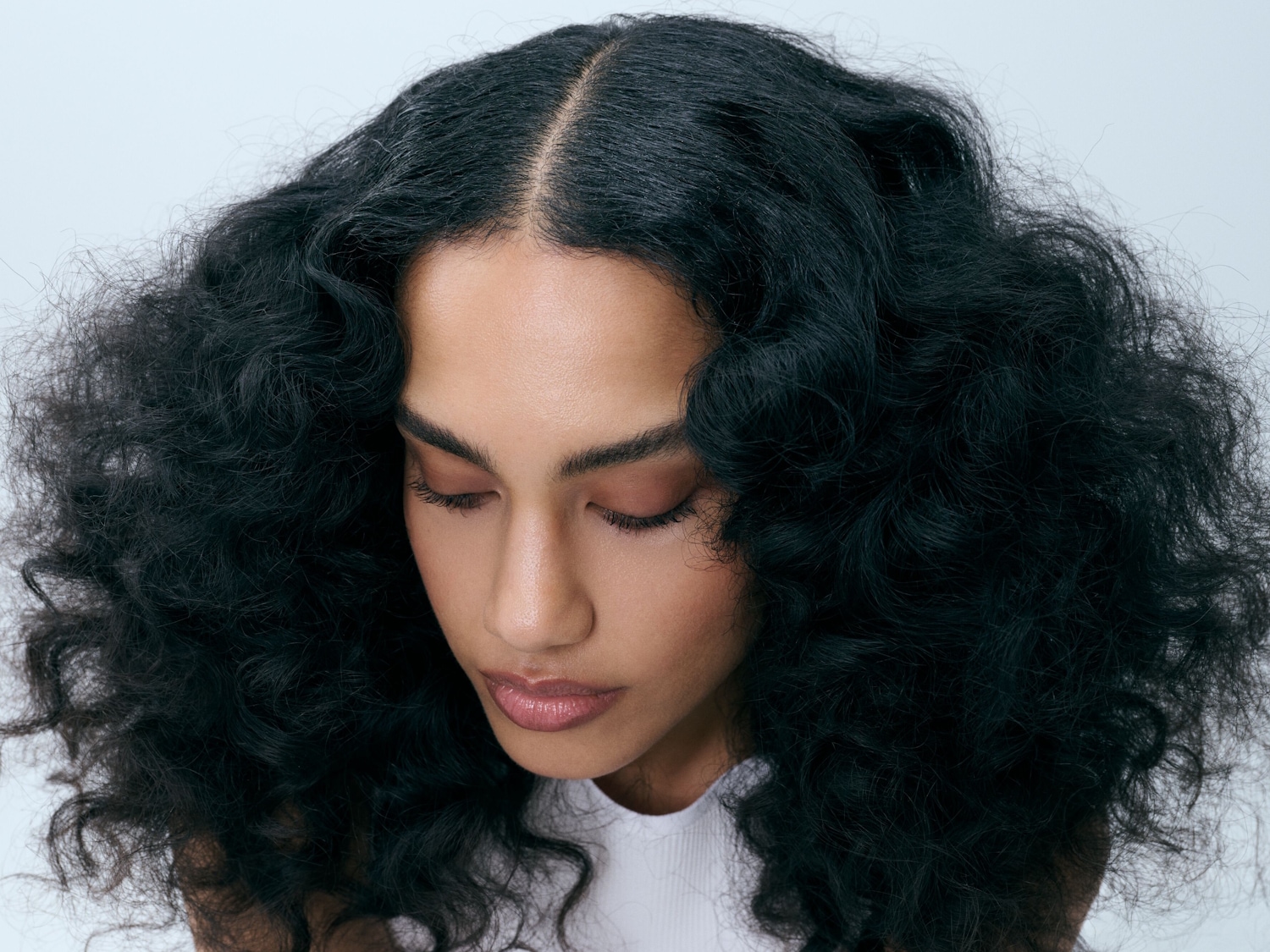 Una donna con lunghi capelli neri ricci e voluminosi, con la riga centrale, indossa un top bianco e ha gli occhi chiusi, mostrando un trucco naturale e labbra lucide.