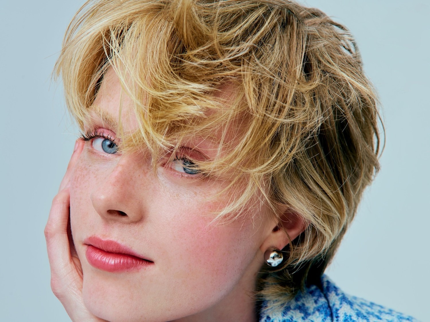 Close-up van een vrouw met blond haar, blauwe ogen en een lichte make-up, die haar gezicht aanraakt en een zilveren oorbel draagt.