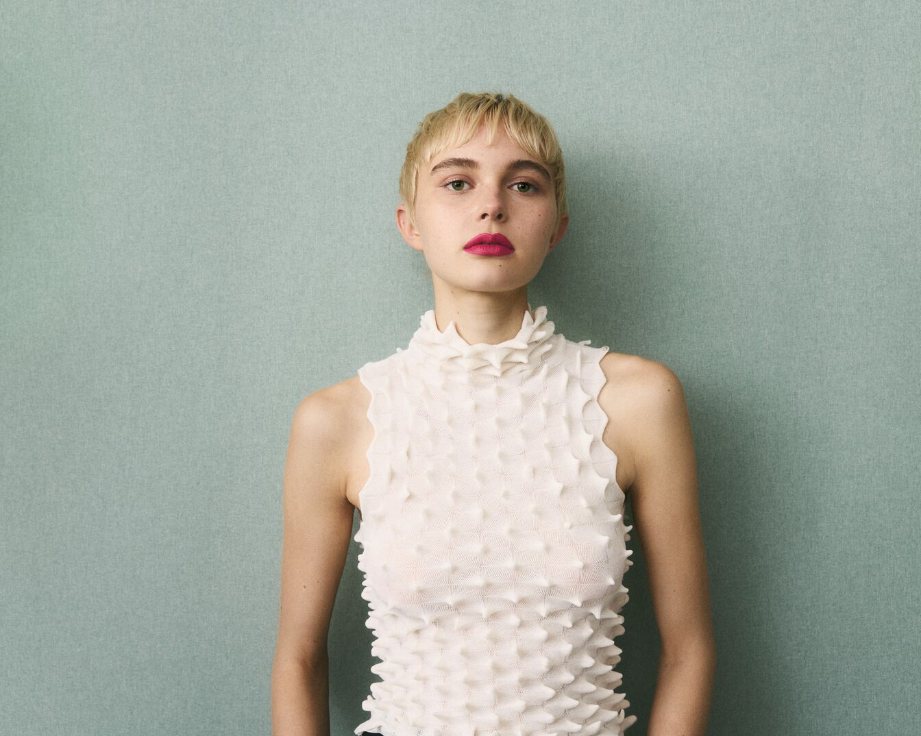 Een jonge vrouw met kort blond haar en roze lippenstift draagt een witte top met een hoge kraag tegen een lichtblauwe achtergrond.