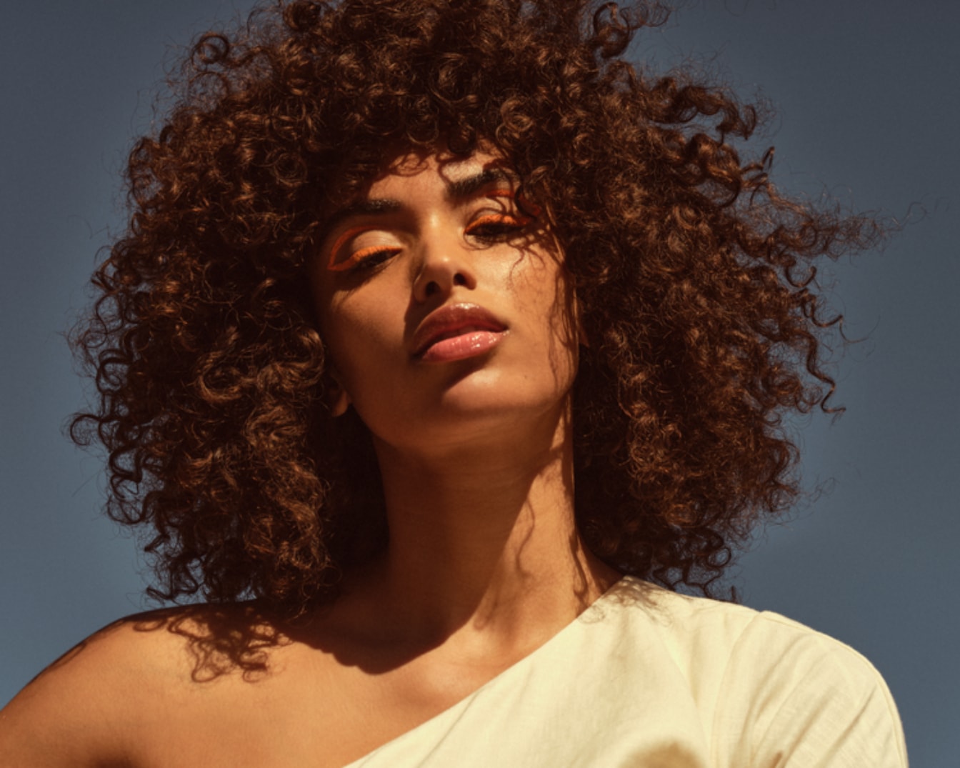 Primo piano di una modella con capelli ricci castani, ombretto arancione brillante e labbra lucide, che mostra un look audace e moderno.