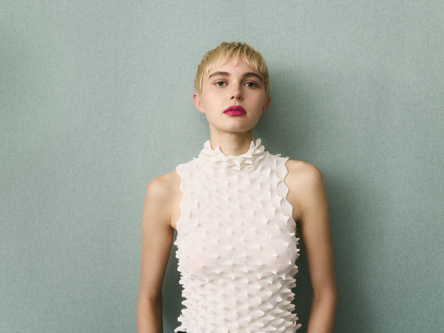 Een jonge vrouw met kort blond haar en roze lippenstift draagt een witte top met een unieke textuur tegen een lichtblauwe achtergrond.