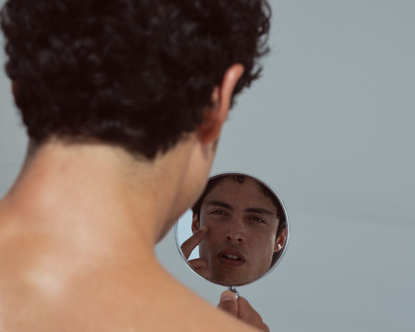 Un uomo con i capelli ricci neri si guarda allo specchio, toccandosi il viso con un dito.