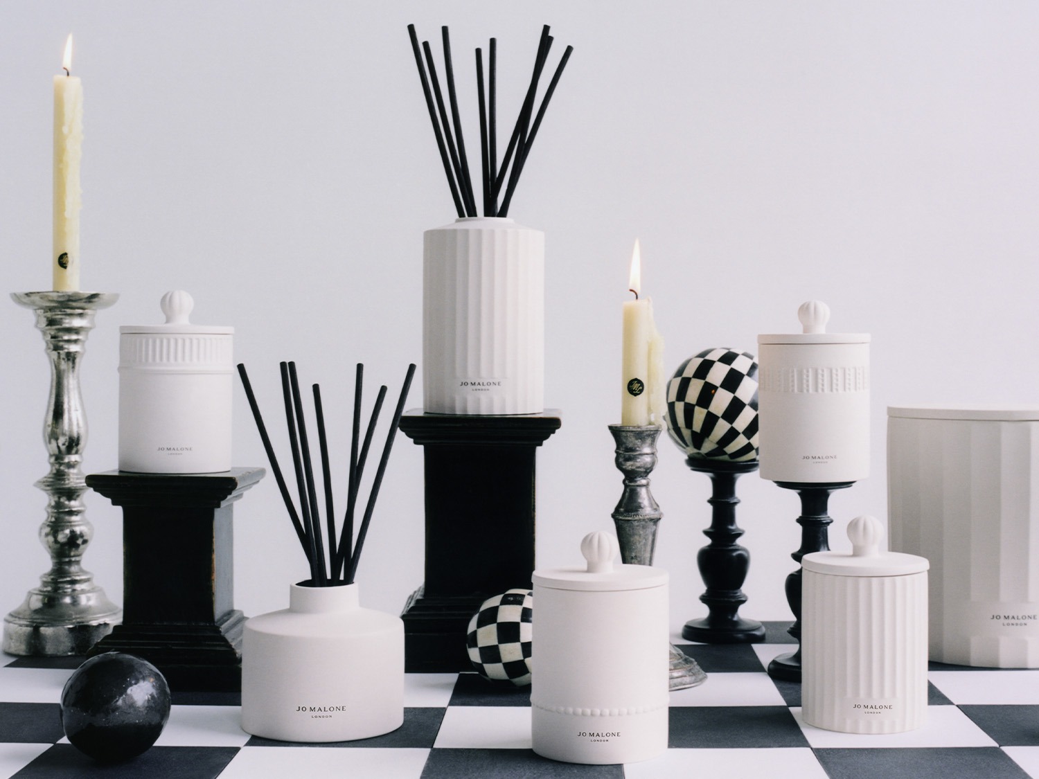 Een verzameling Jo Malone huisgeuren, waaronder diffusers met zwarte stokjes en kaarsen, op een dambordpatroon.