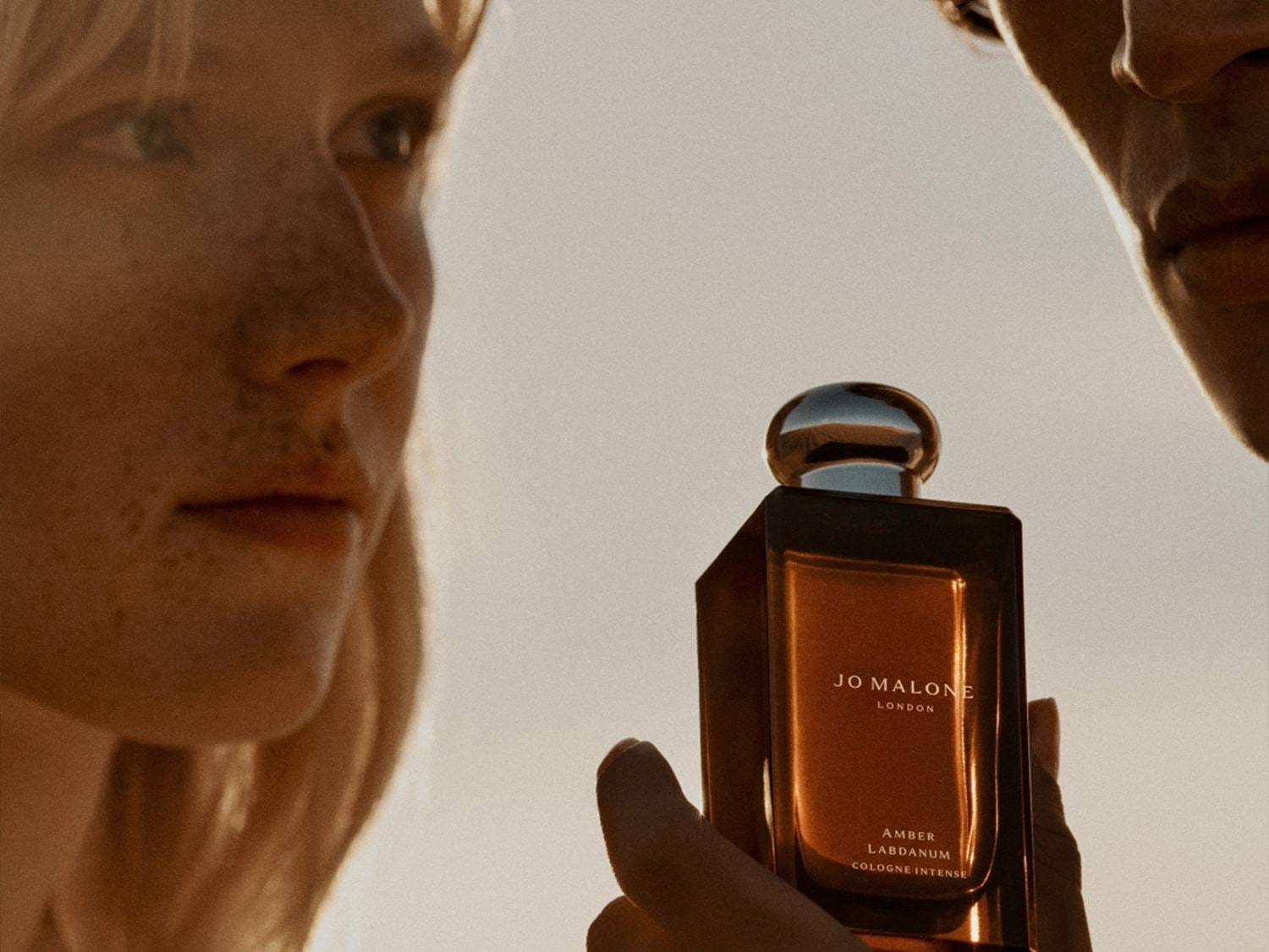 Twee mensen delen een moment met Jo Malone Amber Labdanum Cologne Intense, de amberkleurige vloeistof zichtbaar in de flacon.