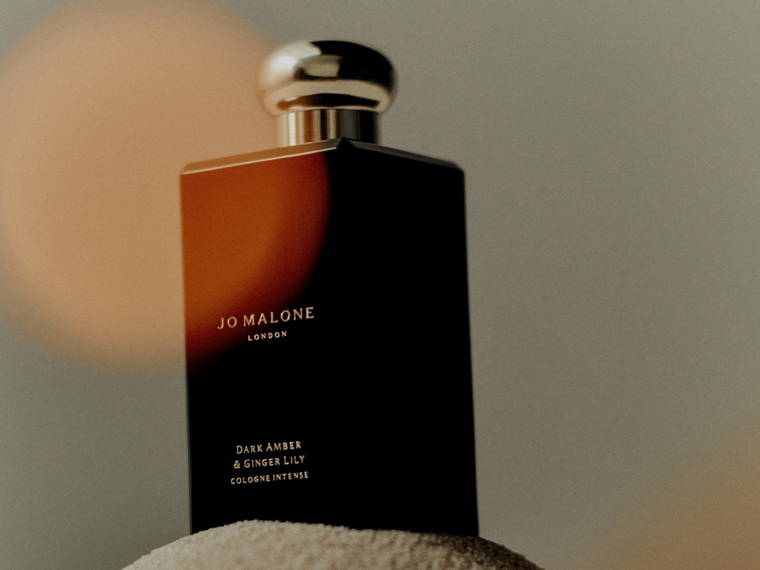 Een rechthoekige, donkerbruine flacon van Jo Malone London, met de tekst 'Dark Amber & Ginger Lily Cologne Intense', staat op een zachte, korrelige ondergrond.