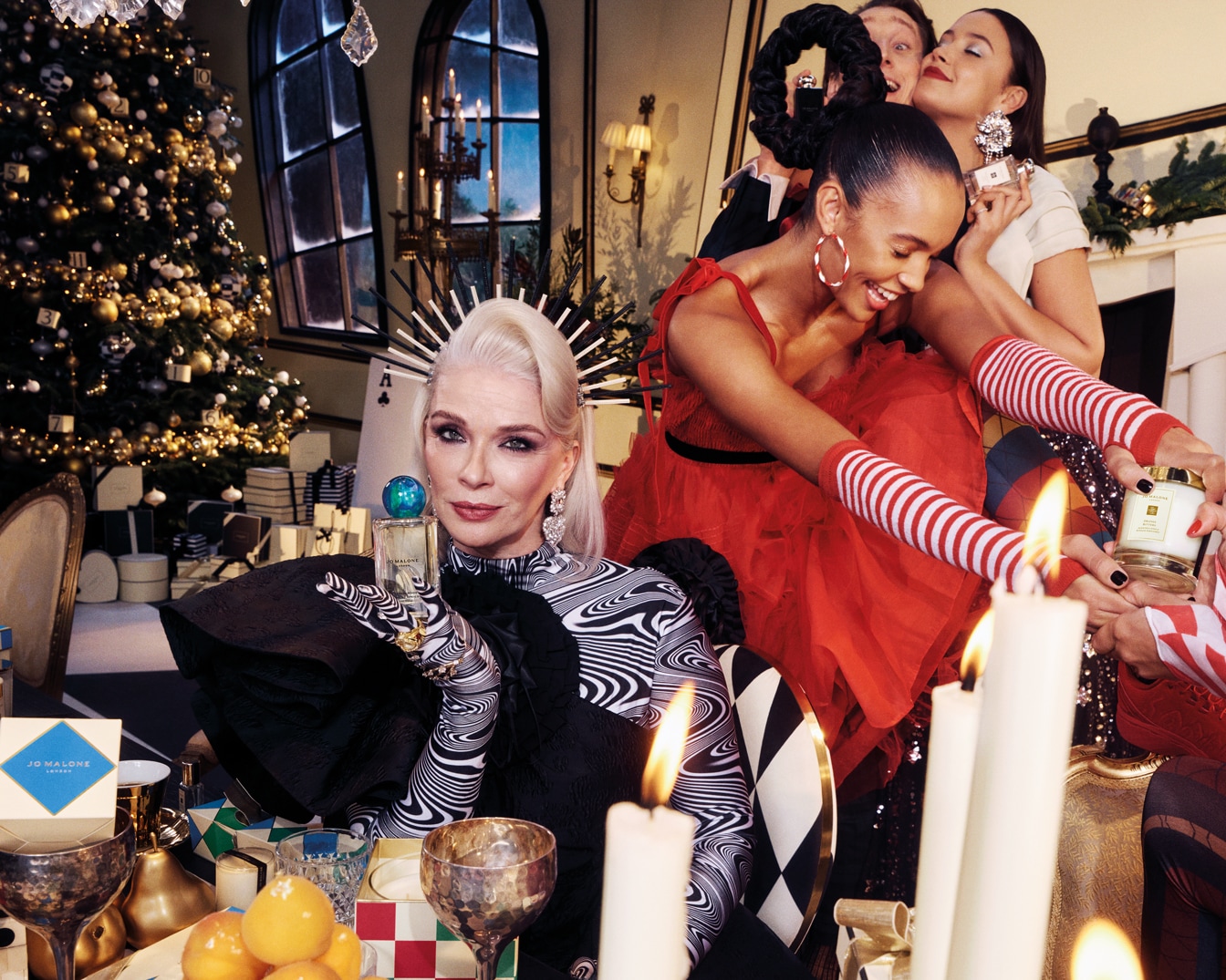 Dans une ambiance festive, une femme élégante en noir et blanc présente un parfum Jo Malone, entourée d'amis et de bougies scintillantes, évoquant la magie des fêtes.