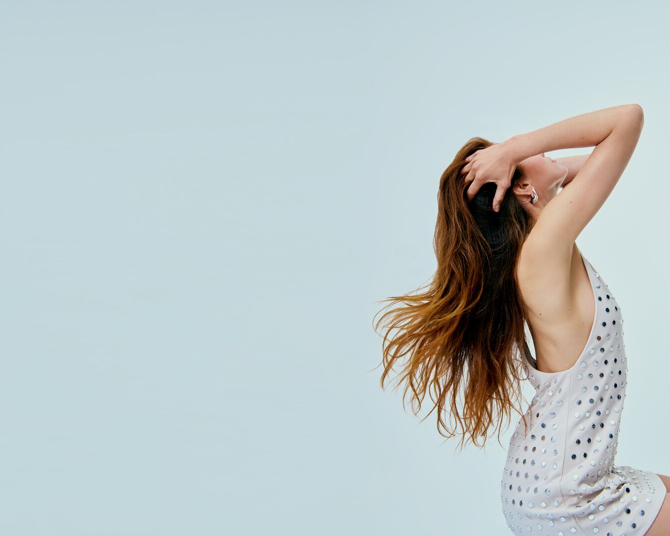Eine Frau mit langen, gewellten, braunen Haaren, die ein ärmelloses, kurzes, helles Kleid mit silbernen Pailletten trägt, posiert mit erhobenen Armen und nach hinten geneigtem Kopf.