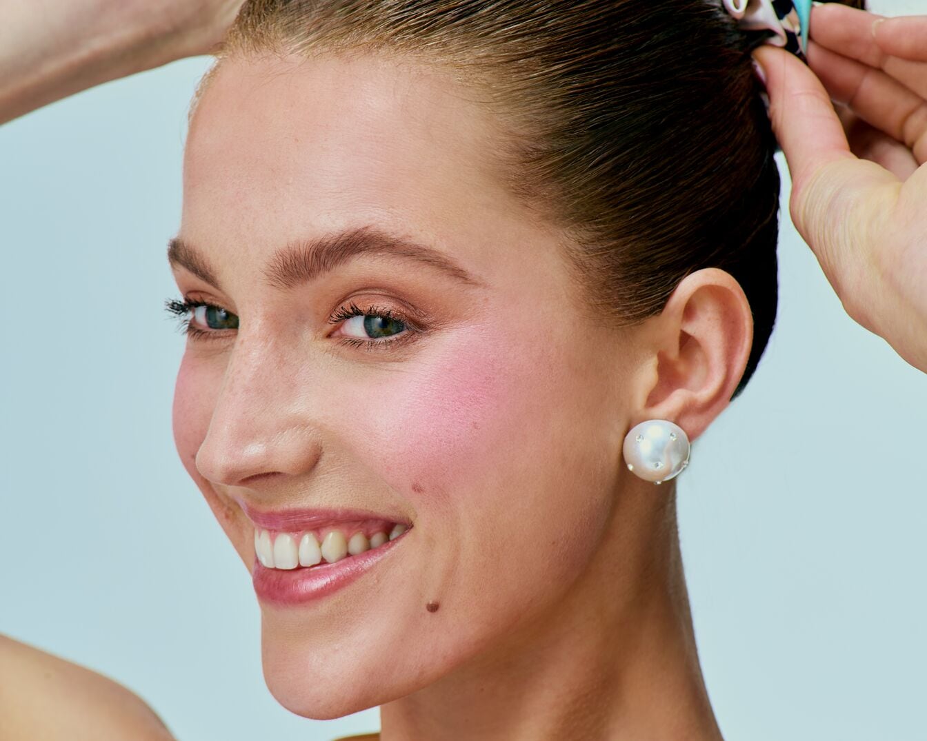 Nahaufnahme einer lächelnden Frau mit rosa Rouge, glänzenden Lippen und einem perlenbesetzten Ohrring, die ihr Haar mit einem bunten Tuch zurückbindet.