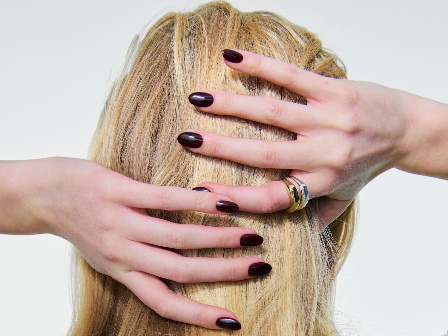 Una mujer rubia se toca el pelo con las manos, luciendo uñas pintadas de color burdeos oscuro y un anillo dorado en el dedo anular.