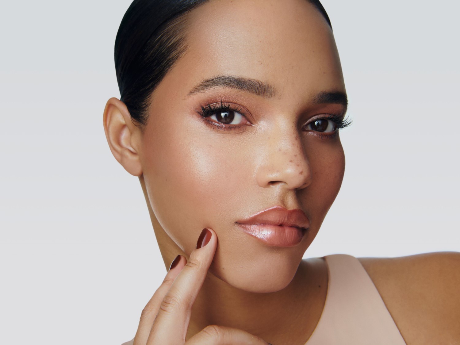 Primer plano de un rostro maquillado con tonos tierra, resaltando los ojos con sombras y pestañas definidas, y los labios con un brillo sutil, mientras una mano toca suavemente la mejilla.