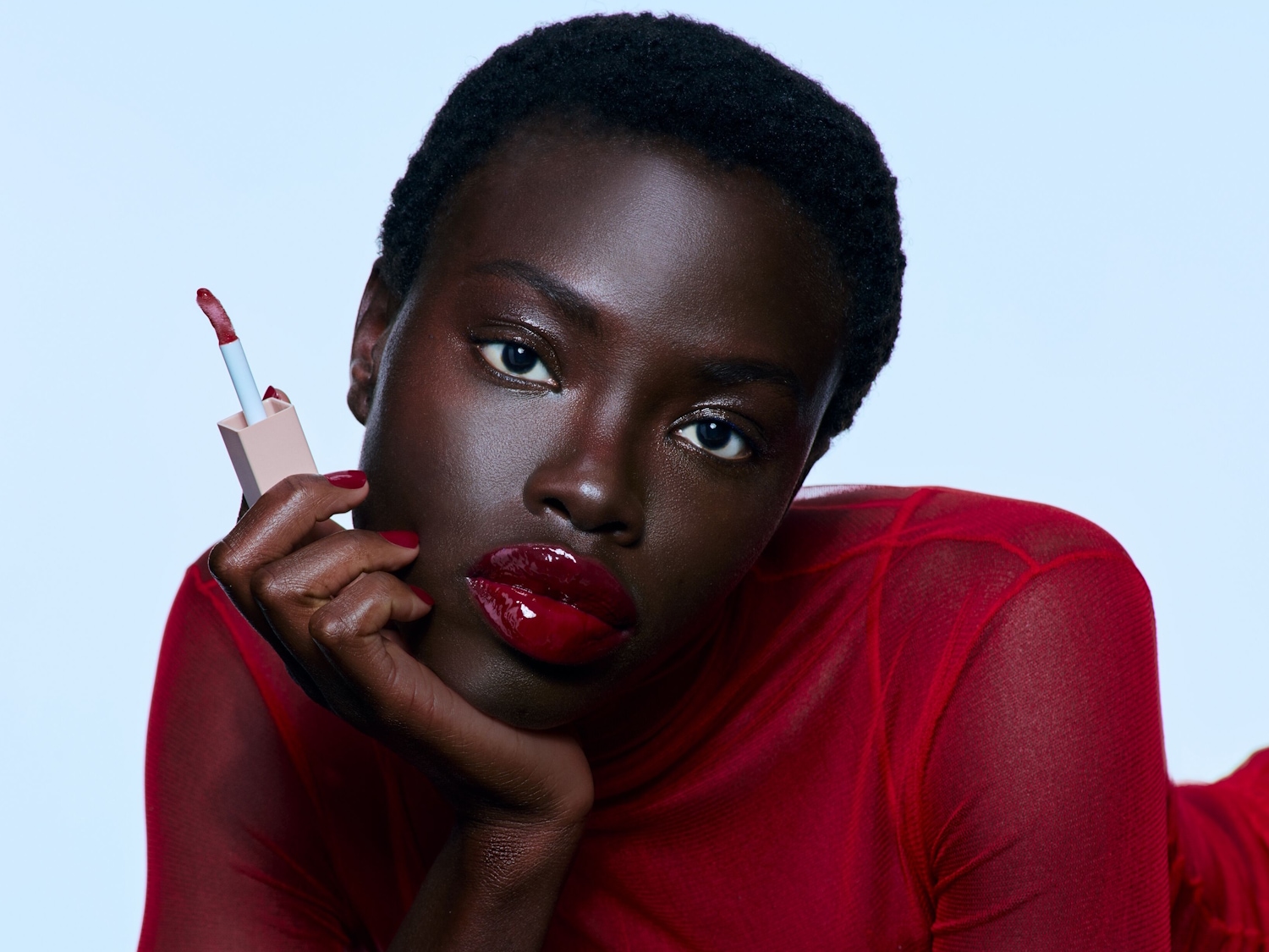 Una modelo de piel oscura luce un brillo de labios rojo intenso, sosteniendo el aplicador con un envase color melocotón, resaltando sus labios brillantes y su top de malla roja.