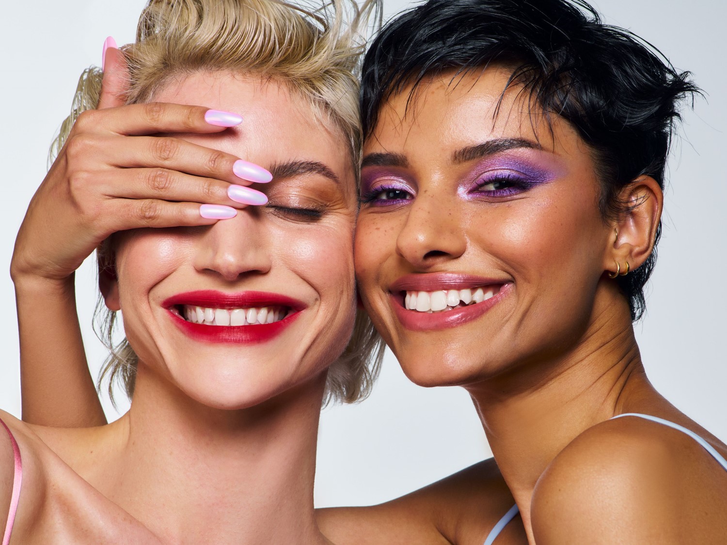 Dos mujeres sonrientes, una con cabello rubio y la otra con cabello oscuro, posan juntas; la rubia tiene los ojos cerrados y una mano con uñas iridiscentes sobre su frente, mientras que la morena luce un vibrante maquillaje de ojos morado.