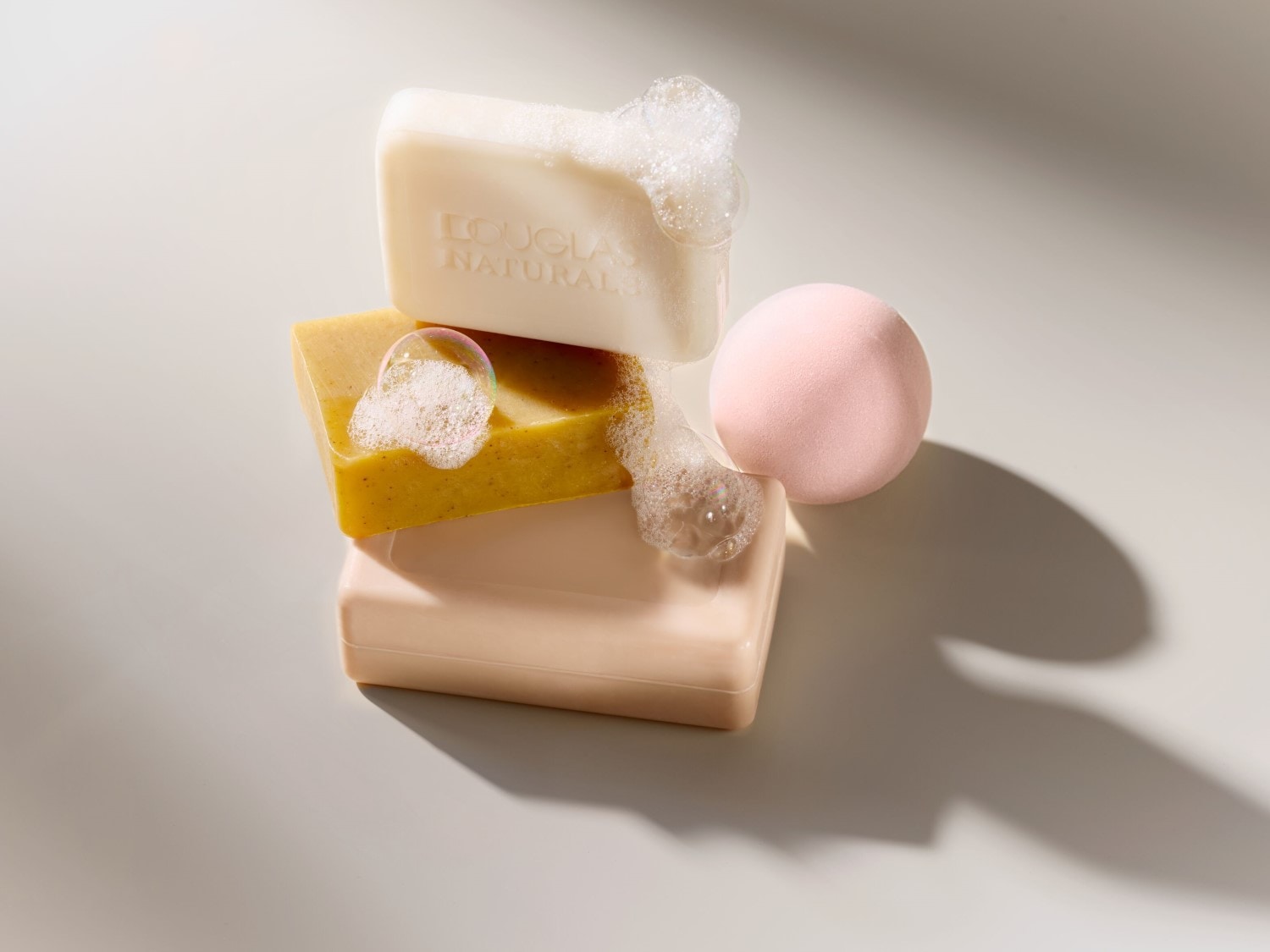 Stapel van drie Douglas Natural zepen in wit, geel en beige, met schuim en een roze make-up spons.