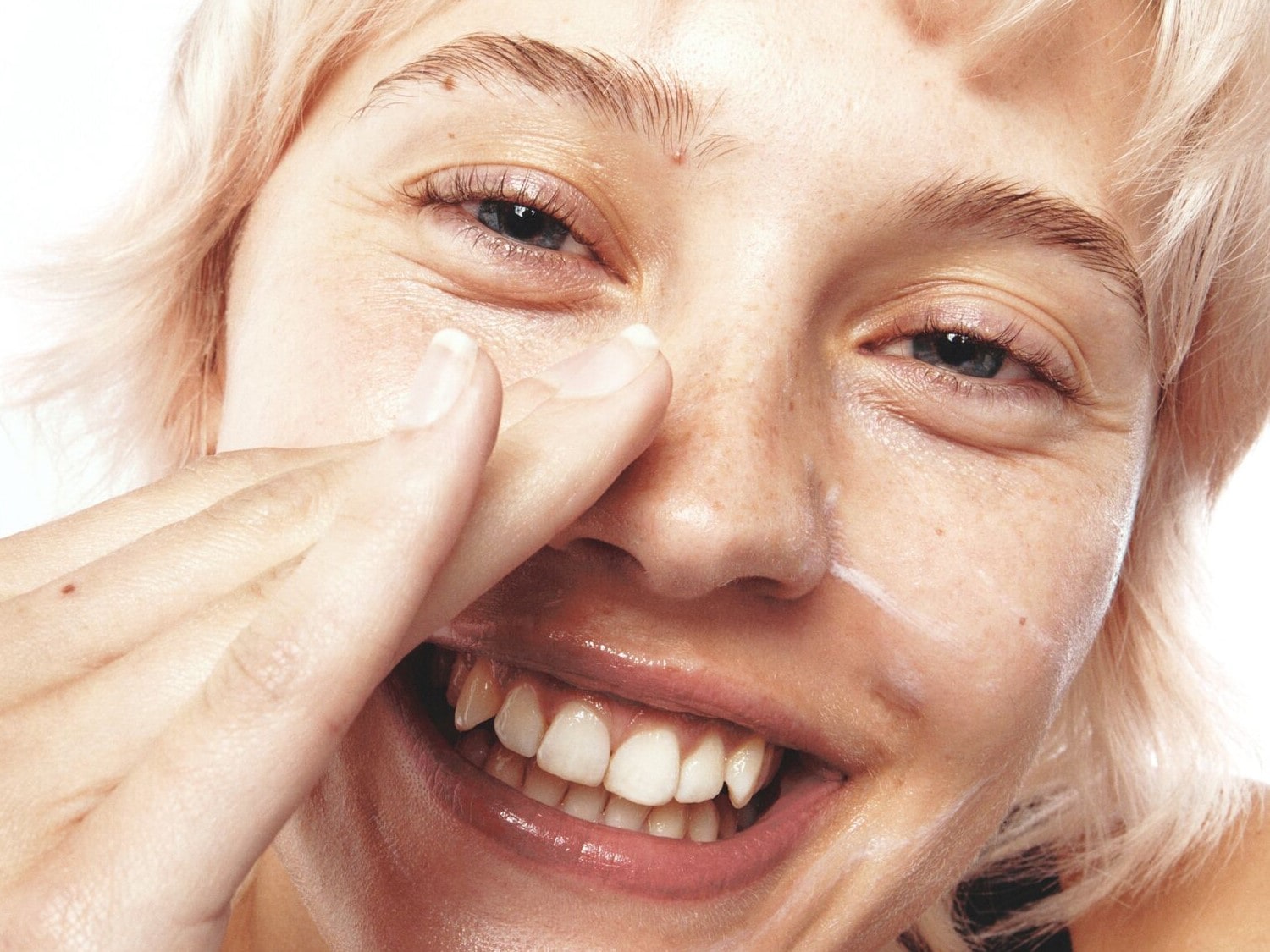 Een close-up van een lachende vrouw met blond haar die een witte crème aanbrengt op haar gezicht, waarbij haar vingers haar neus aanraken.