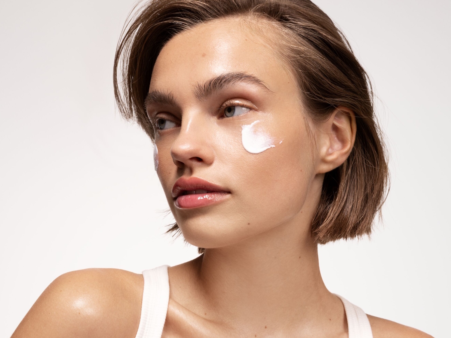 Nahaufnahme einer Frau mit kurzen braunen Haaren und einem weißen Top, die eine weiße Creme auf ihrer Wange aufträgt, um ihre Haut zu pflegen.