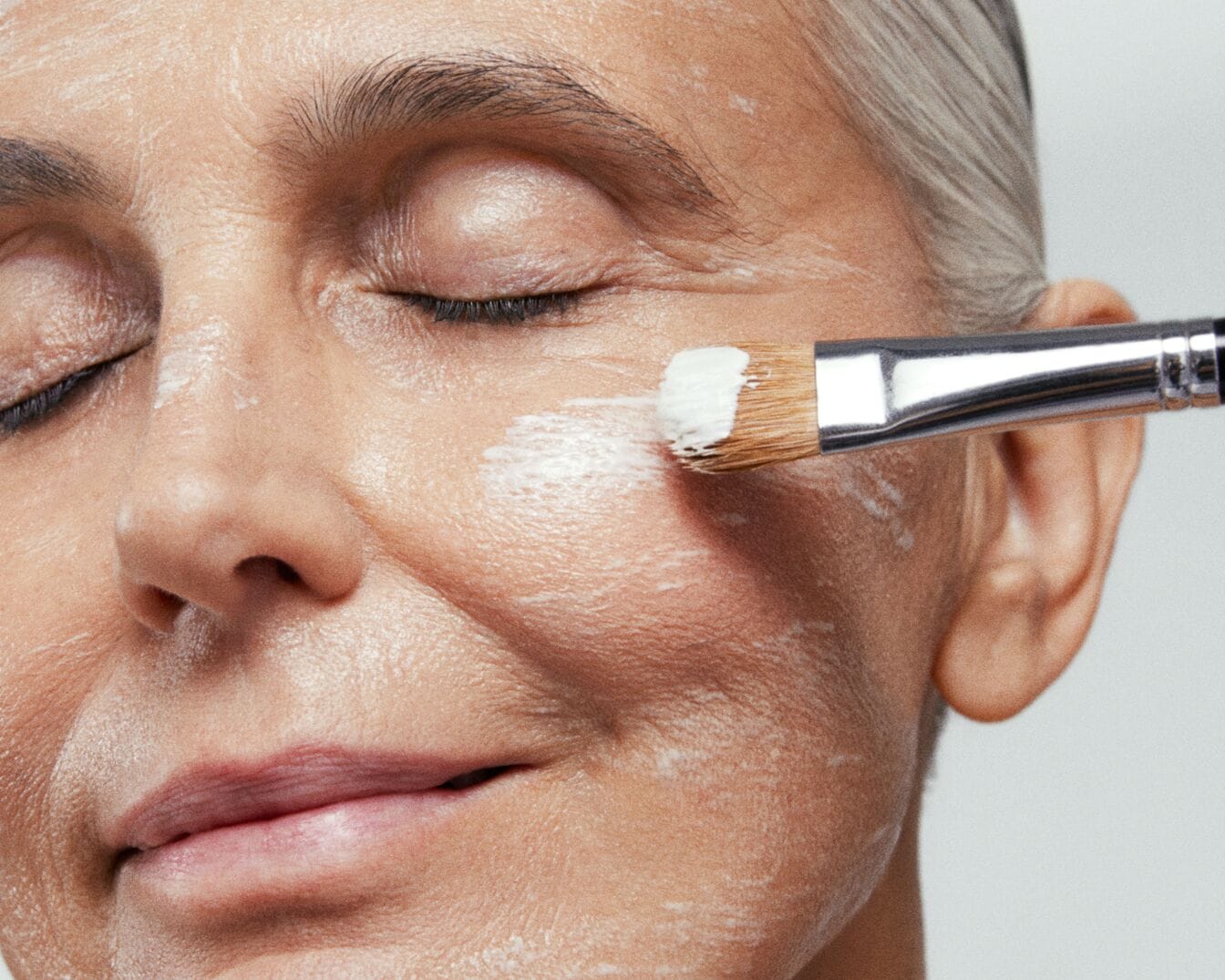 Con los ojos cerrados, una mujer de piel clara se aplica una mascarilla blanca con una brocha plateada para un cutis radiante.