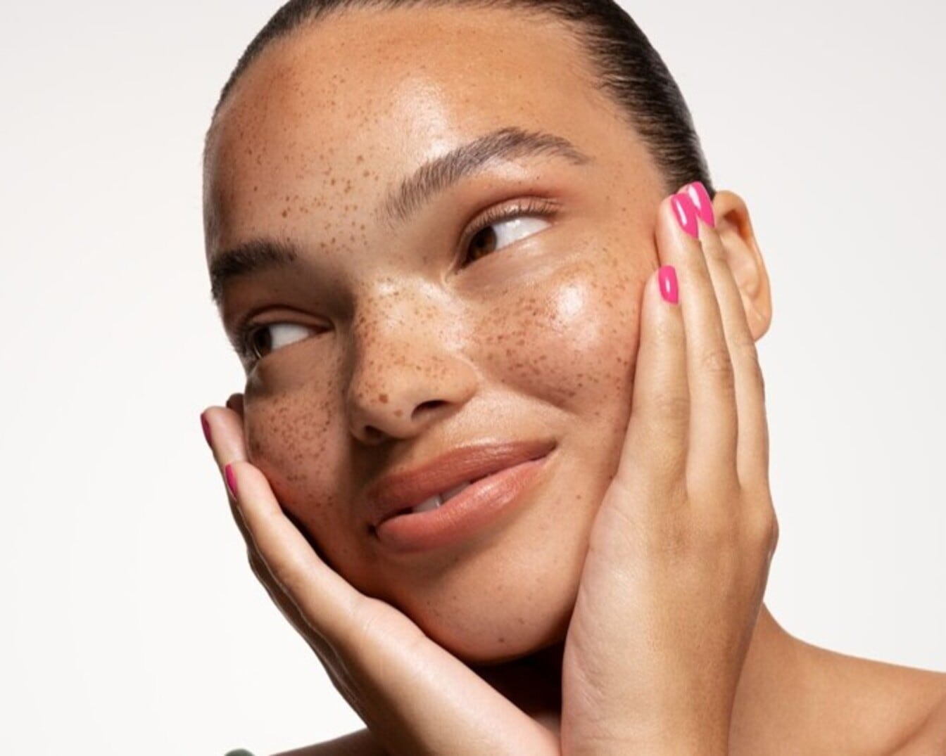 Primer plano de una mujer sonriente con pecas y uñas rosas, tocándose la cara con las manos, resaltando su piel radiante y su expresión alegre.