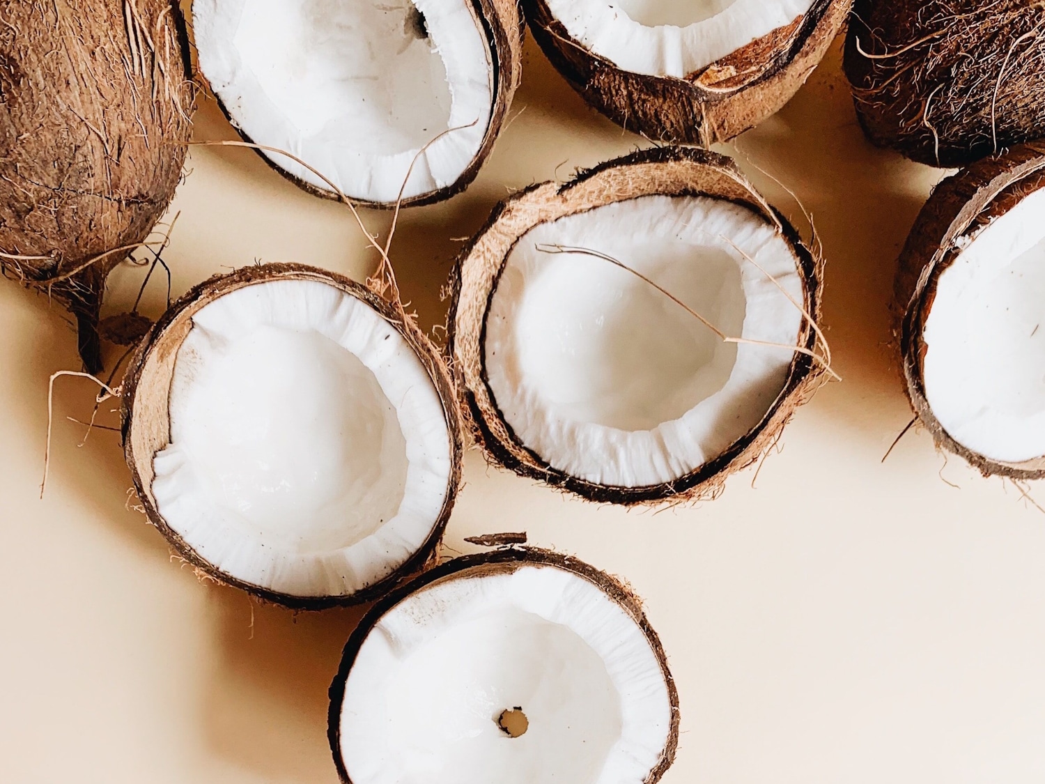 Primer plano de varios cocos, algunos enteros con cáscara marrón y otros partidos por la mitad, mostrando la pulpa blanca sobre un fondo beige.