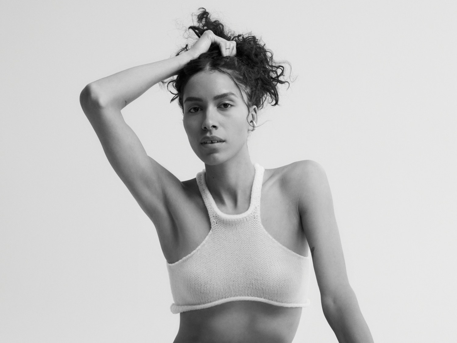 Schwarz-Weiß-Aufnahme einer Frau mit lockigem Haar, die ein weißes Strickoberteil trägt und eine Hand im Haar hat.