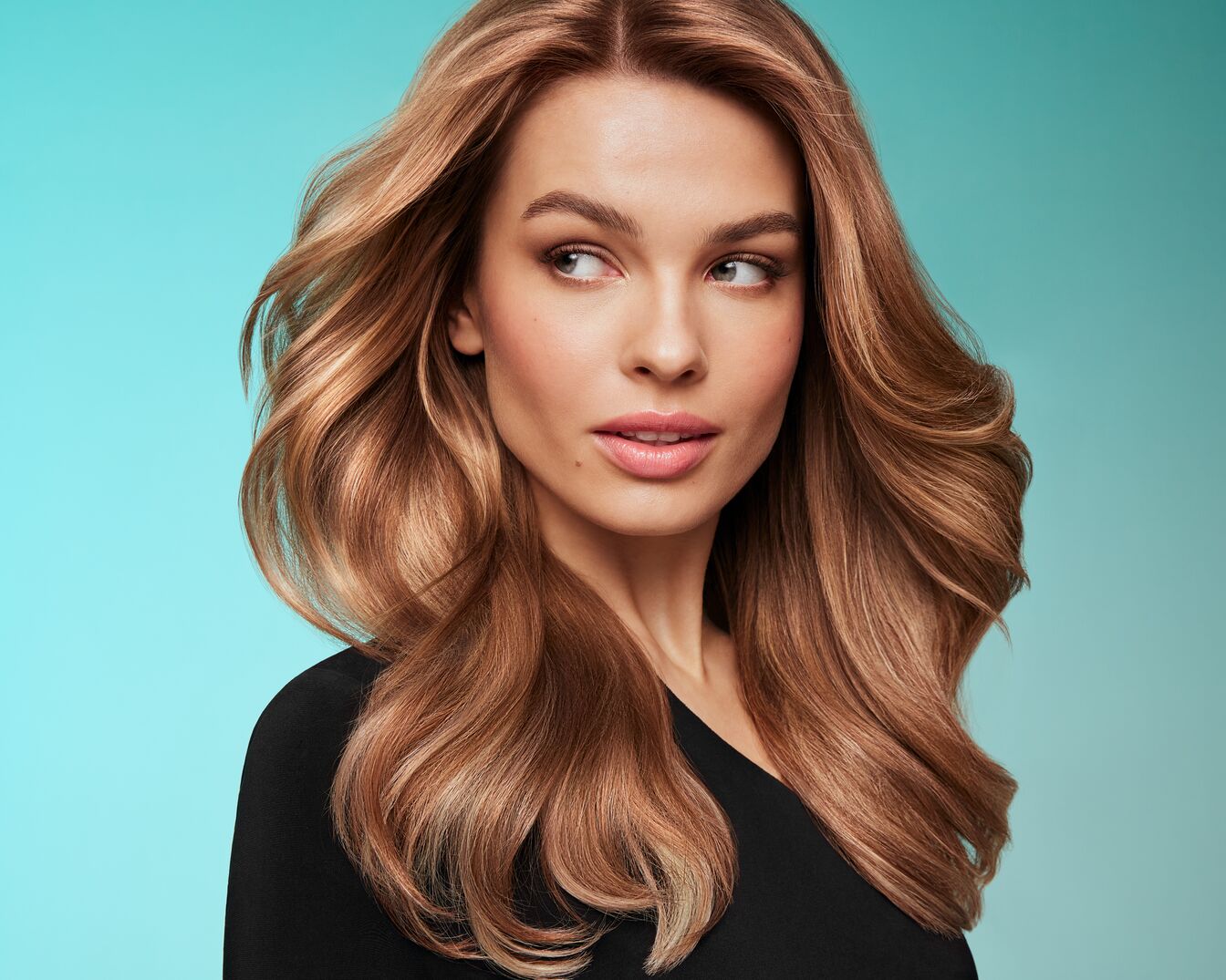 Portret kobiety z długimi, falowanymi włosami w odcieniach blondu i brązu, ubrana w czarną bluzkę na tle błękitnego gradientu.