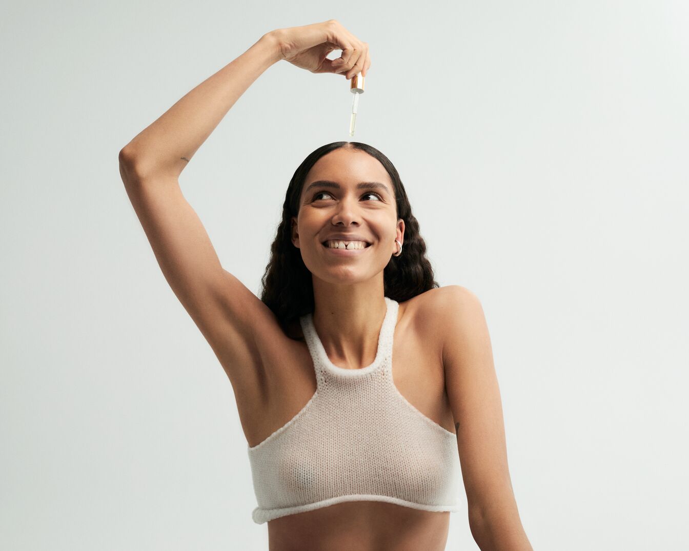 Una donna sorridente applica un siero con un contagocce sui capelli, indossando un top bianco lavorato a maglia, per una cura di bellezza che nutre e illumina.
