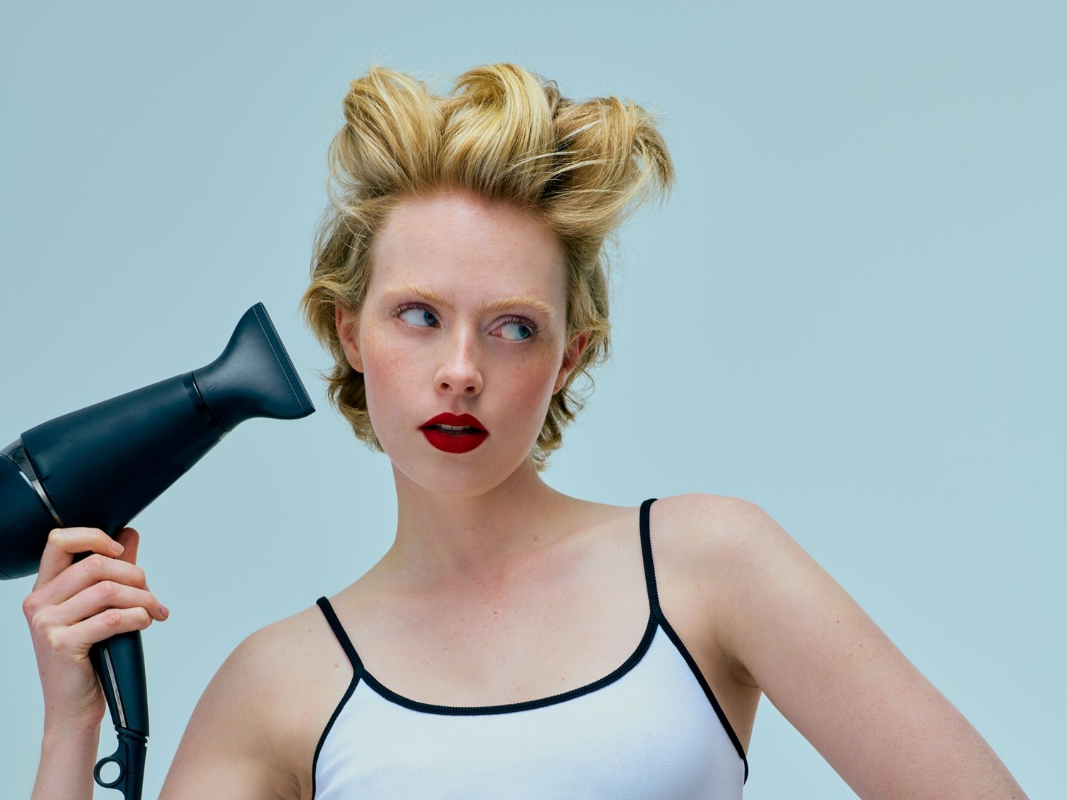 Una donna bionda con labbra rosse e un top bianco con spalline nere tiene un asciugacapelli nero, guardando di lato con i capelli acconciati in riccioli voluminosi.