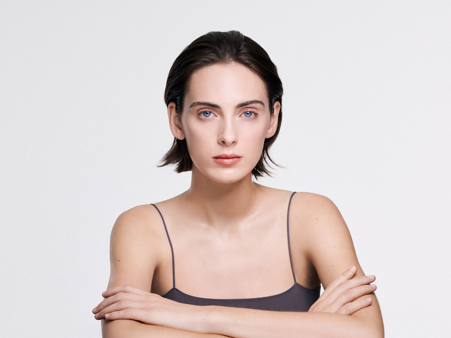 Porträt einer Frau mit kurzen, dunklen Haaren und blauen Augen, die ein graues Top trägt und die Arme vor der Brust verschränkt.