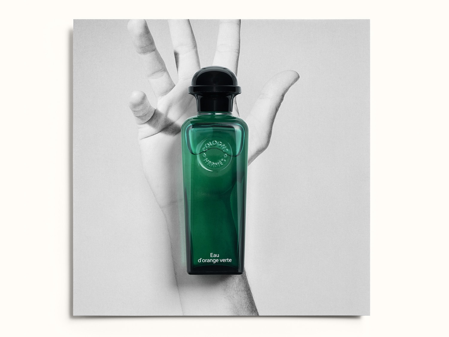 Una bottiglia di profumo verde smeraldo con tappo nero, "Eau d'Orange Verte" di Hermès, è tenuta da una mano in bianco e nero, che ne esalta il colore vibrante.