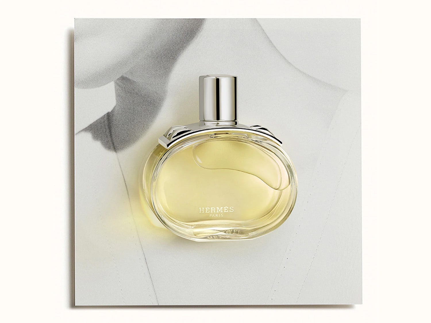 Un flacone di profumo Hermès rotondo e trasparente con liquido giallo dorato e tappo argentato è posizionato su uno sfondo bianco e grigio che ricorda un disegno astratto.