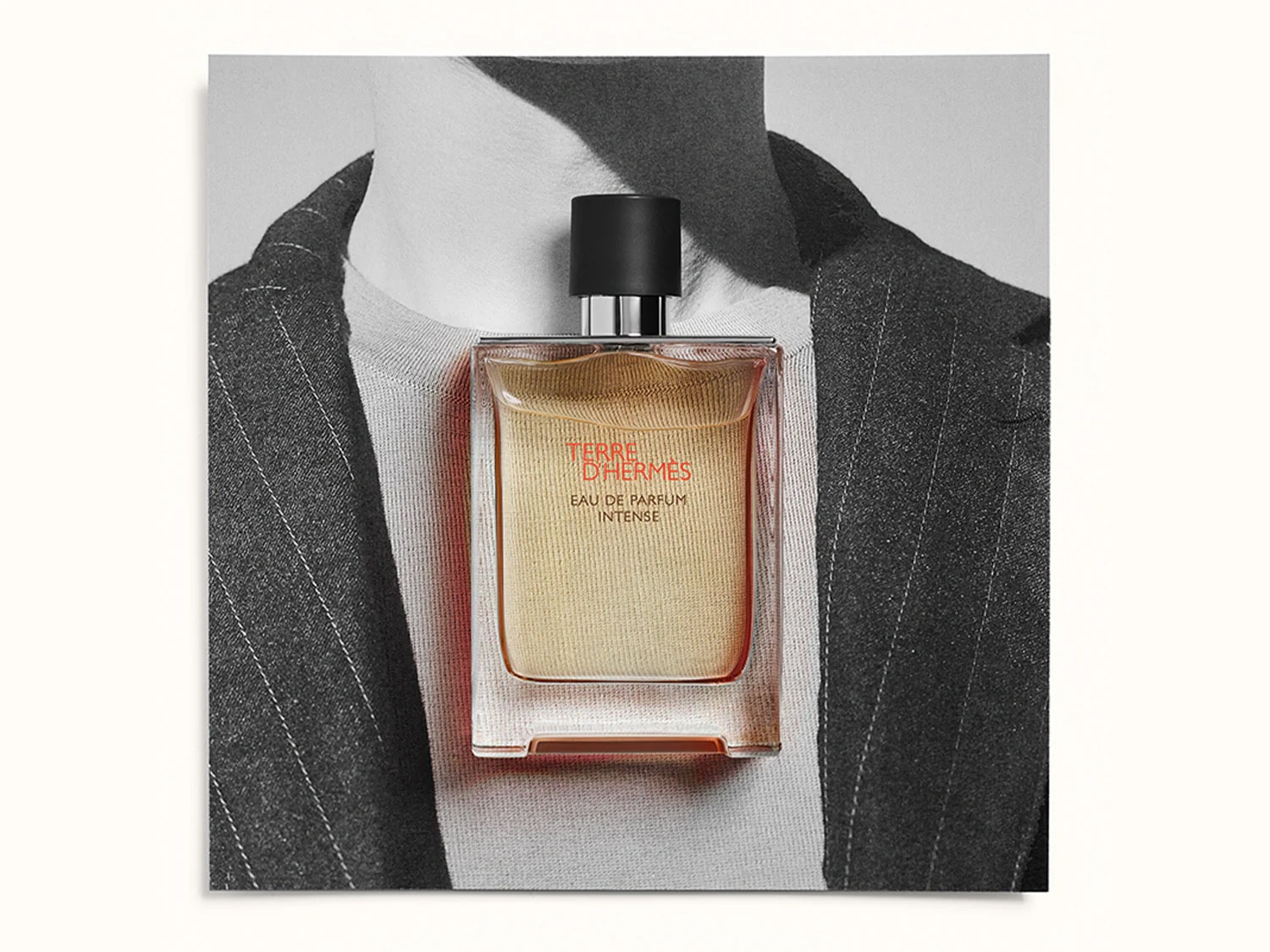 Un flacone rettangolare di Terre d'Hermès Eau de Parfum Intense, con liquido ambrato e tappo nero, è posizionato su uno sfondo che mostra il collo di una persona con una giacca a righe.