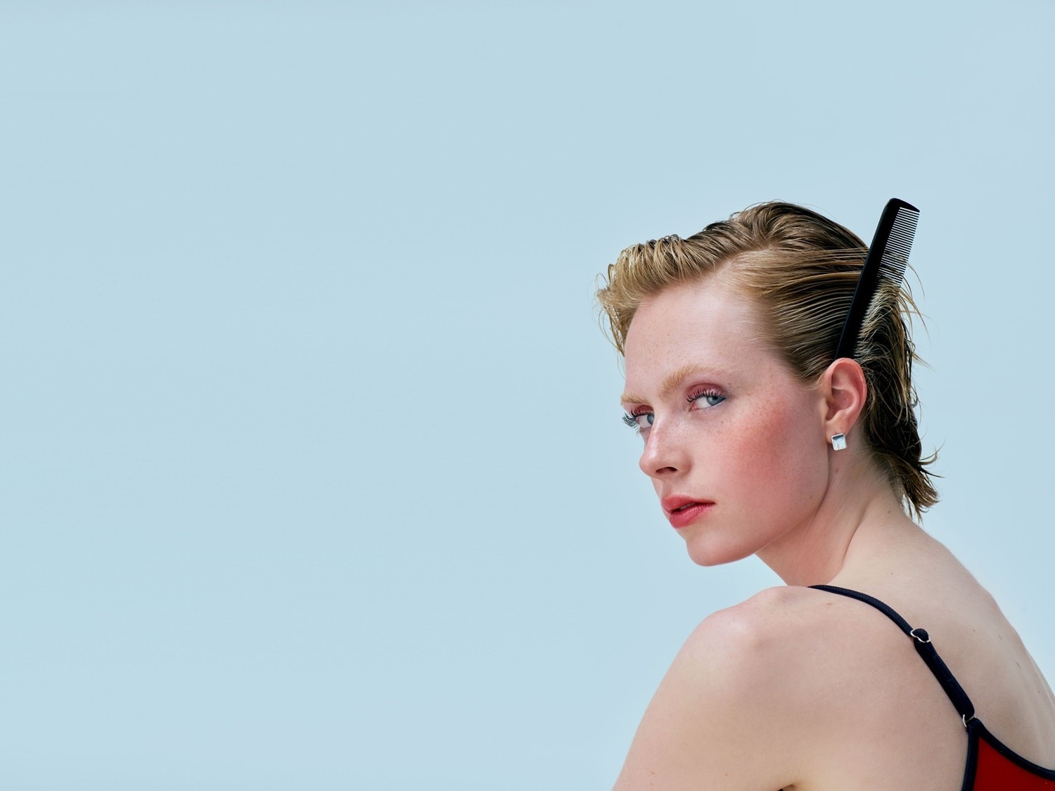 Een vrouw met kort, blond haar en een zwarte kam erin, kijkt over haar schouder met rode lippenstift en blush tegen een lichtblauwe achtergrond.