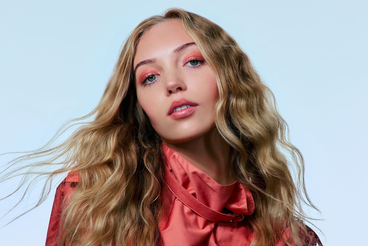 Porträt einer jungen Frau mit gewelltem, blondem Haar, einem korallfarbenen Oberteil und passendem Lidschatten, der ihren grünen Augen schmeichelt.