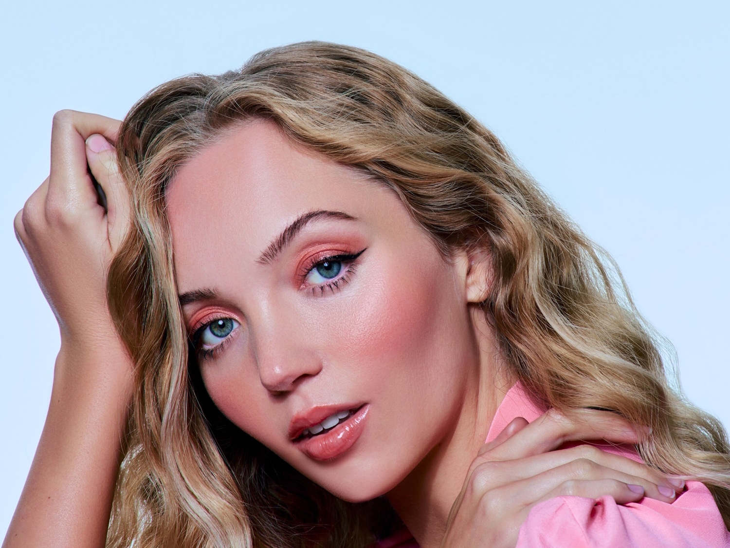 Nahaufnahme einer blonden Frau mit gewelltem Haar, die ein rosa Outfit trägt und ein komplettes Make-up mit rosa Lidschatten und glänzenden Lippen hat.