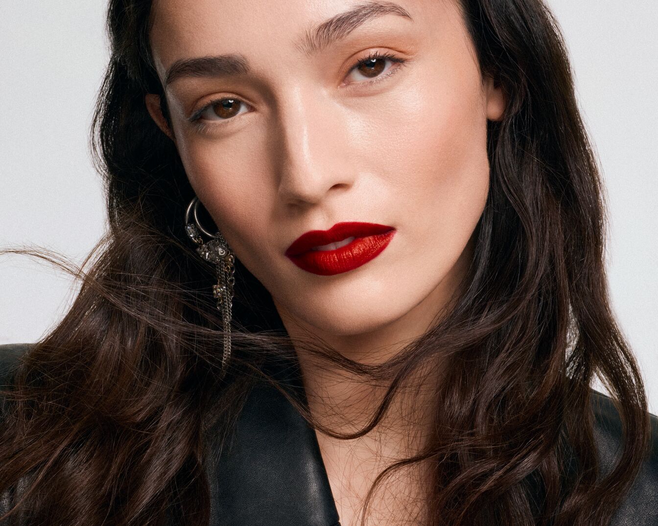Nahaufnahme einer Frau mit welligem, dunklem Haar, die einen roten Lippenstift und einen silbernen Ohrring mit baumelnden Ketten trägt.