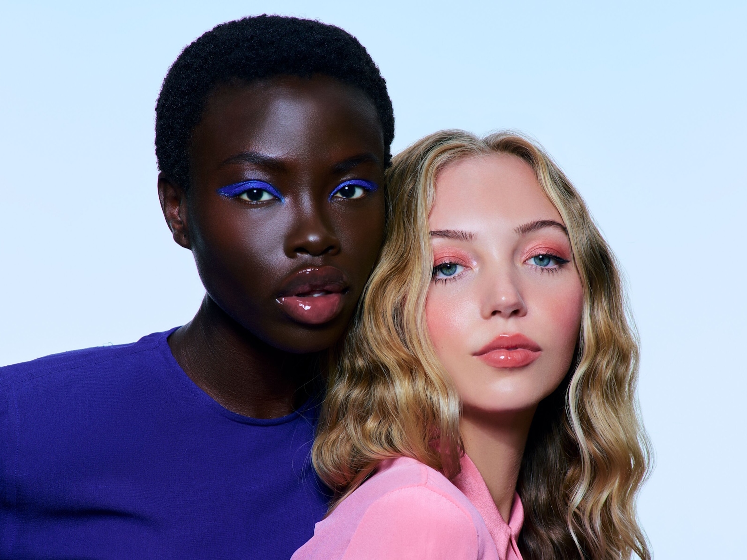 Nahaufnahme von zwei Models: Eine mit blauem Eyeliner und lila Oberteil, die andere mit rosa Lidschatten und rosa Oberteil.