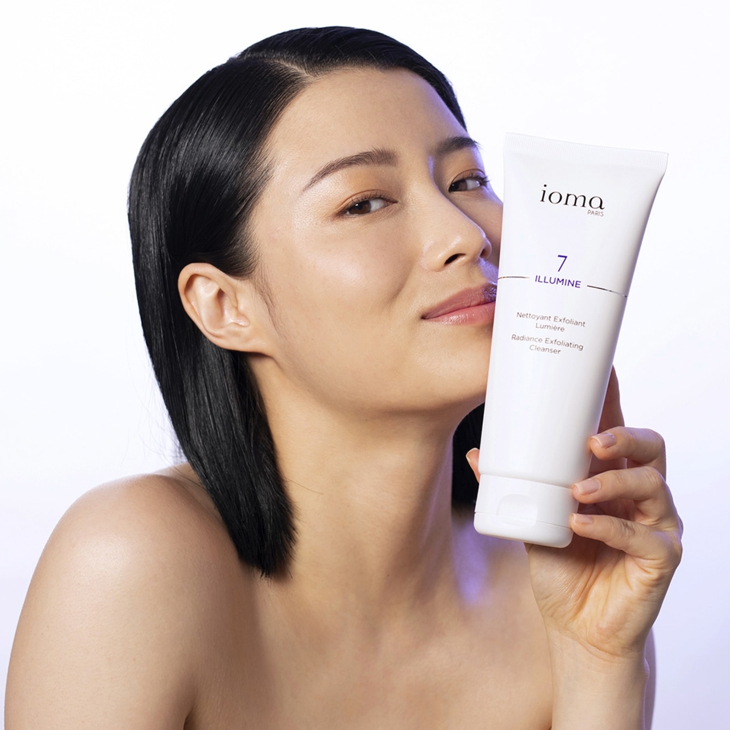 Una donna asiatica tiene in mano un tubo bianco di Ioma Paris 7 Illumine, un detergente esfoliante radiante per una pelle luminosa.