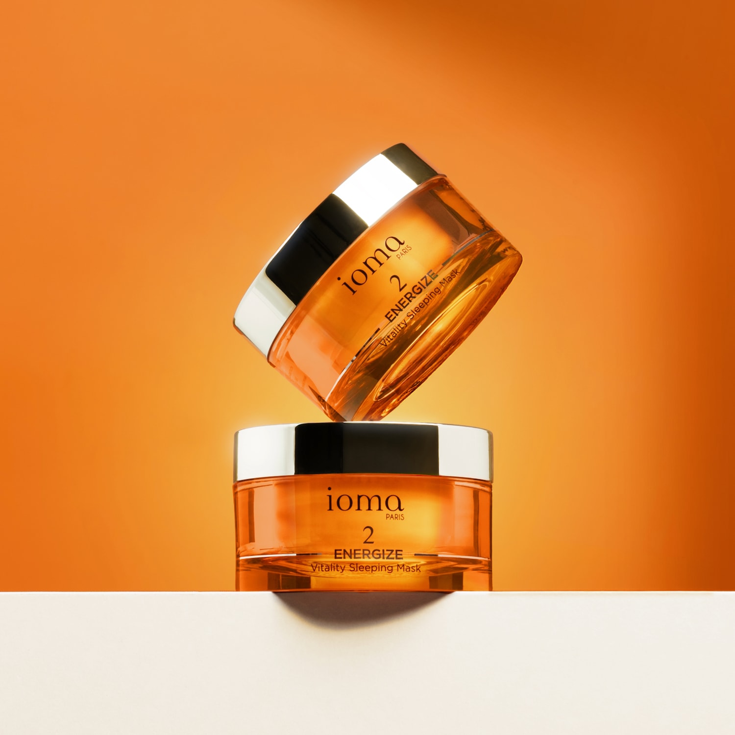 Su uno sfondo arancione, due barattoli impilati di maschera da notte Ioma Paris Energize Vitality, con confezione trasparente e tappo argento.