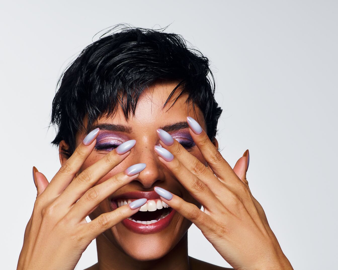 Eine strahlende Frau mit lila Lidschatten und hellblau lackierten, mandelförmigen Nägeln bedeckt ihr Gesicht mit den Händen und lächelt.