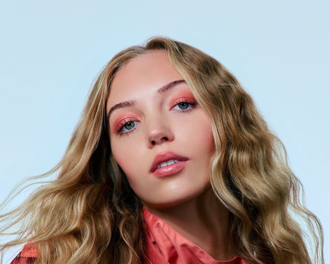 Nahaufnahme eines Models mit gewelltem, blondem Haar, das ein korallenrotes Augen-Make-up und einen passenden Lippenstift trägt, vor einem hellblauen Hintergrund.