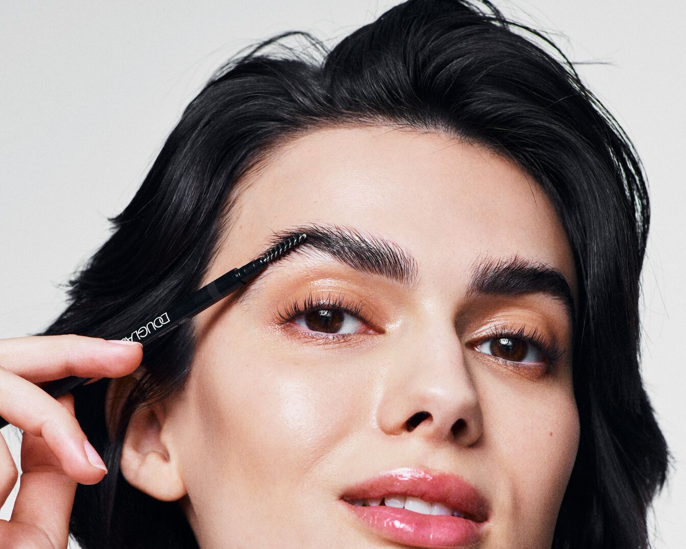Een vrouw met donker haar gebruikt een Douglas wenkbrauwborstel om haar wenkbrauwen te perfectioneren, voor een verzorgde en gedefinieerde look.