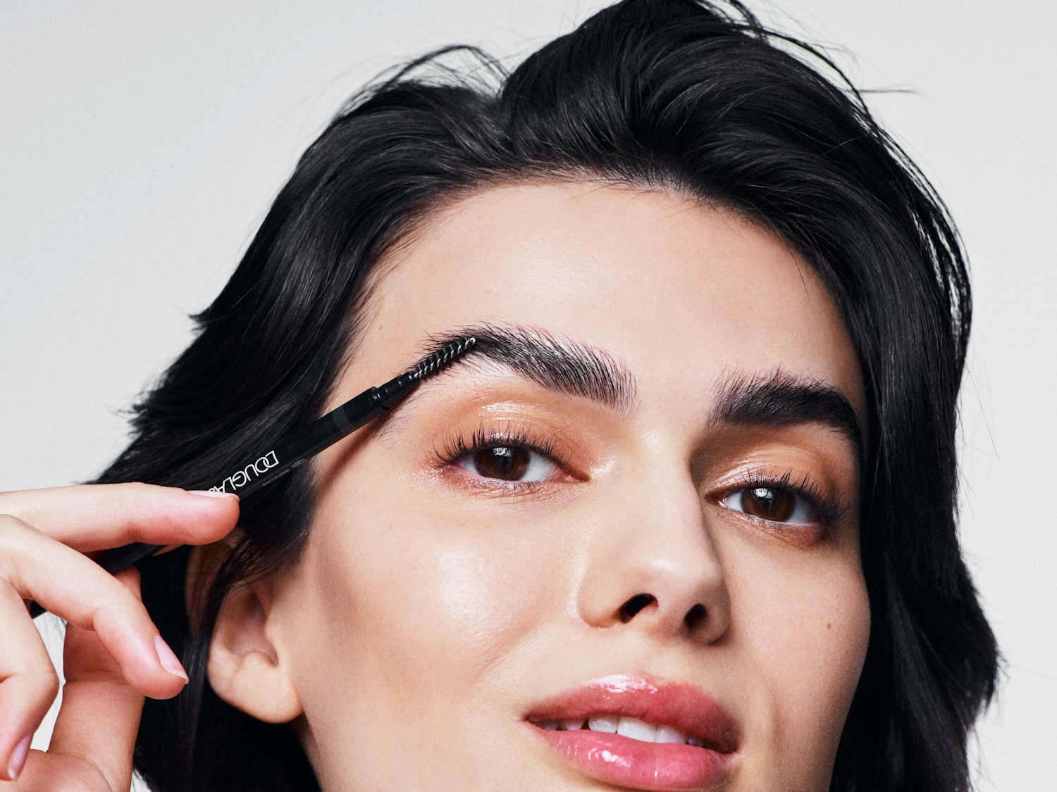 Een vrouw met donker haar gebruikt een Douglas wenkbrauwborstel om haar wenkbrauwen te perfectioneren, voor een verzorgde en gedefinieerde look.