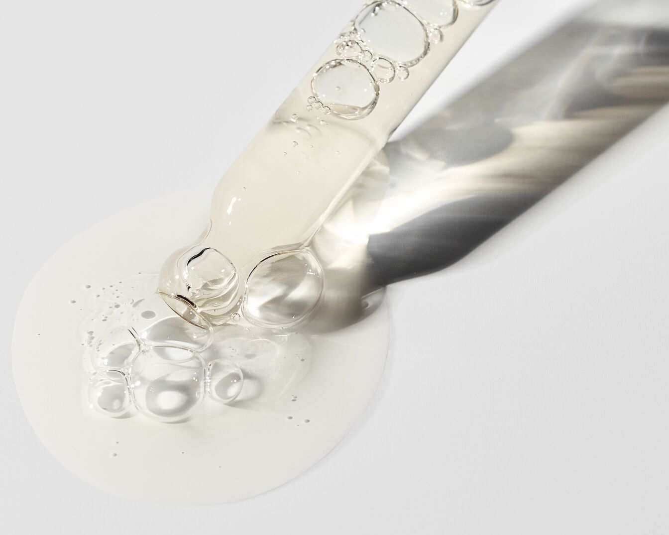 Nahaufnahme einer Pipette mit Serum, das auf eine weiße Oberfläche tropft und eine klare, leicht gelbliche Flüssigkeit mit Bläschen zeigt.