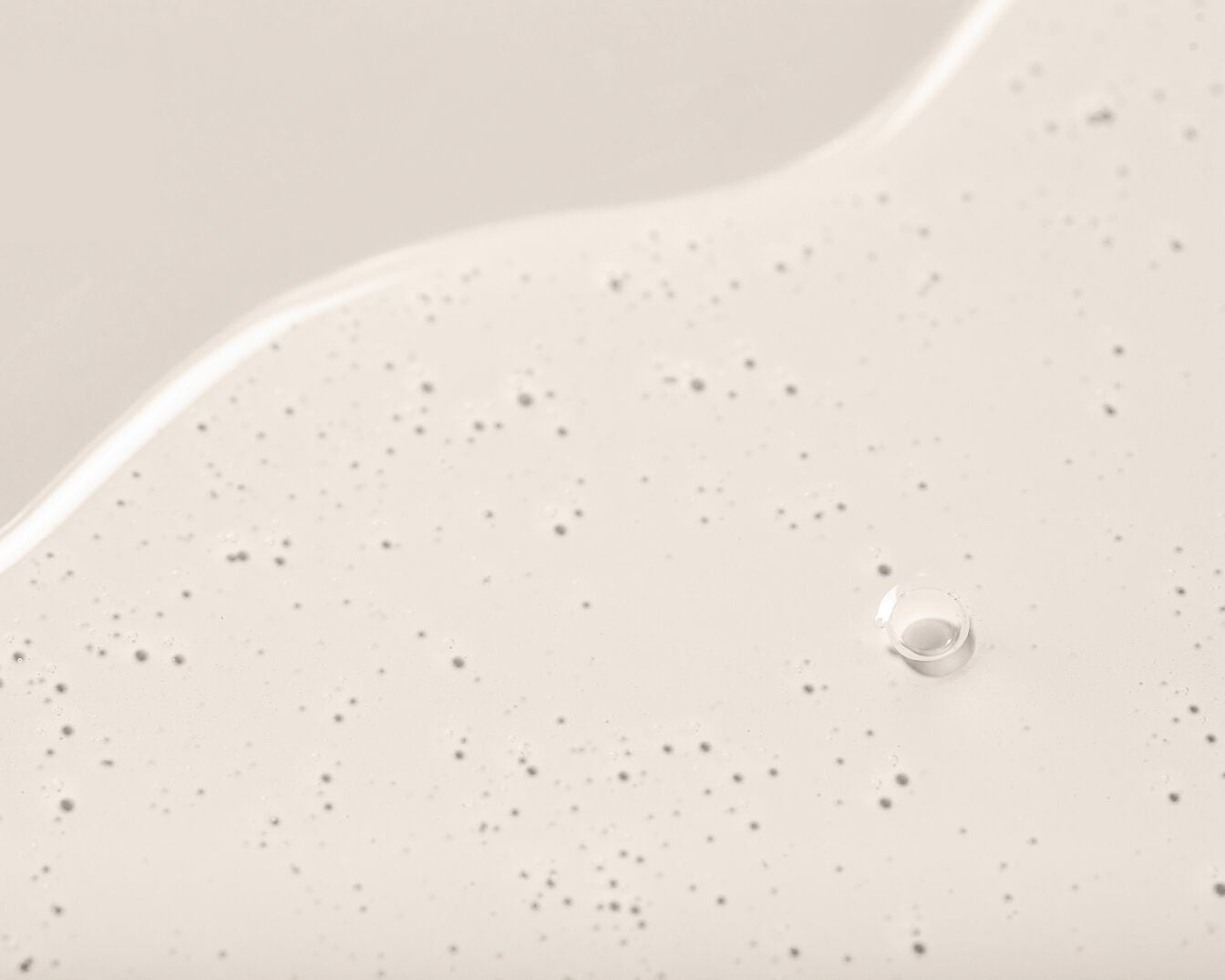 Nahaufnahme einer beigen Flüssigkeit mit kleinen Bläschen und einem einzelnen klaren Tropfen, die eine glatte, leicht reflektierende Textur zeigt.