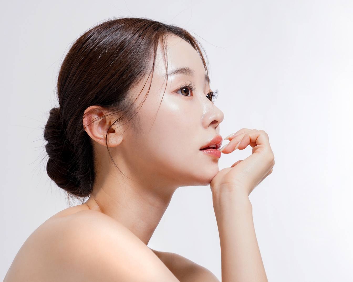 Profil einer Frau mit glänzender Haut, die ihr Gesicht mit der Hand berührt, während sie mit einem Dutt und nackten Schultern posiert.